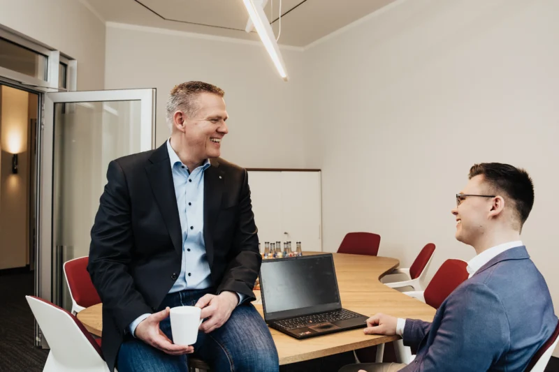 Mann mit Kaffetasse sitzt auf Schreibtisch vor ihm eine junger Mann mit Laptop
